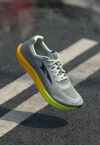 Αθλητικό παπούτσι τρεξίματος με γκρι ανοιχτό πλέγμα, λευκά κορδόνια και κίτρινη βαθμιαία σόλα, αιωρούμενο πάνω από οδόστρωμα με βαμμένες γραμμές.