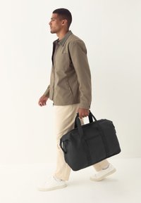 Mustast nailonist duffelkott kahe ülemise sangaga, lukuga sektsioonide ja esitaskuga. Modell kannab beeži jopet, heleid pükse ja tenniseid.