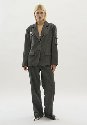 Ensemble de costume gris à motifs comprenant un blazer structuré avec des poches à rabat et un pantalon légèrement ample assorti, complété par des chaussures métalliques à bout pointu.