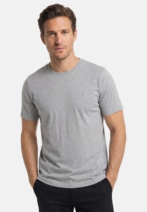 Uomo con capelli castani corti che indossa una t-shirt a maniche corte di colore grigio chiaro semplice e pantaloni neri, in piedi con le mani nelle tasche.