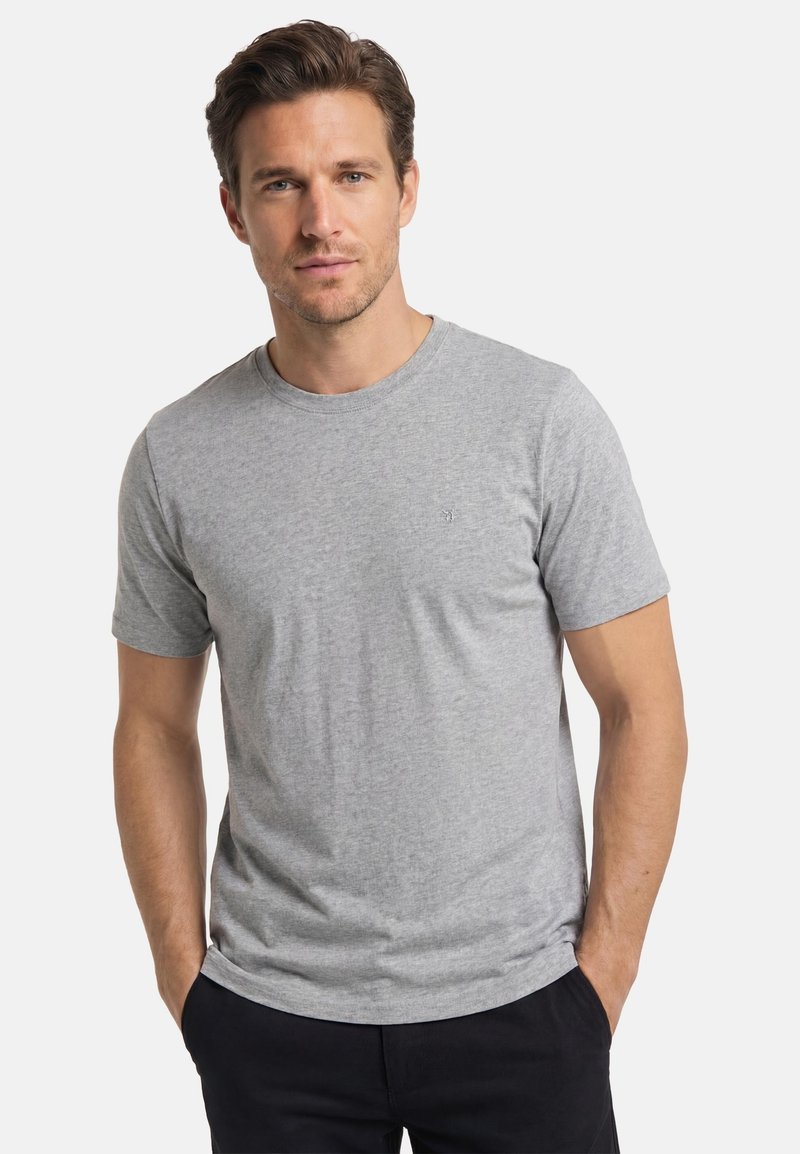 Homme aux cheveux bruns courts portant un t-shirt gris clair uni à manches courtes et un pantalon noir, debout les mains dans les poches.
