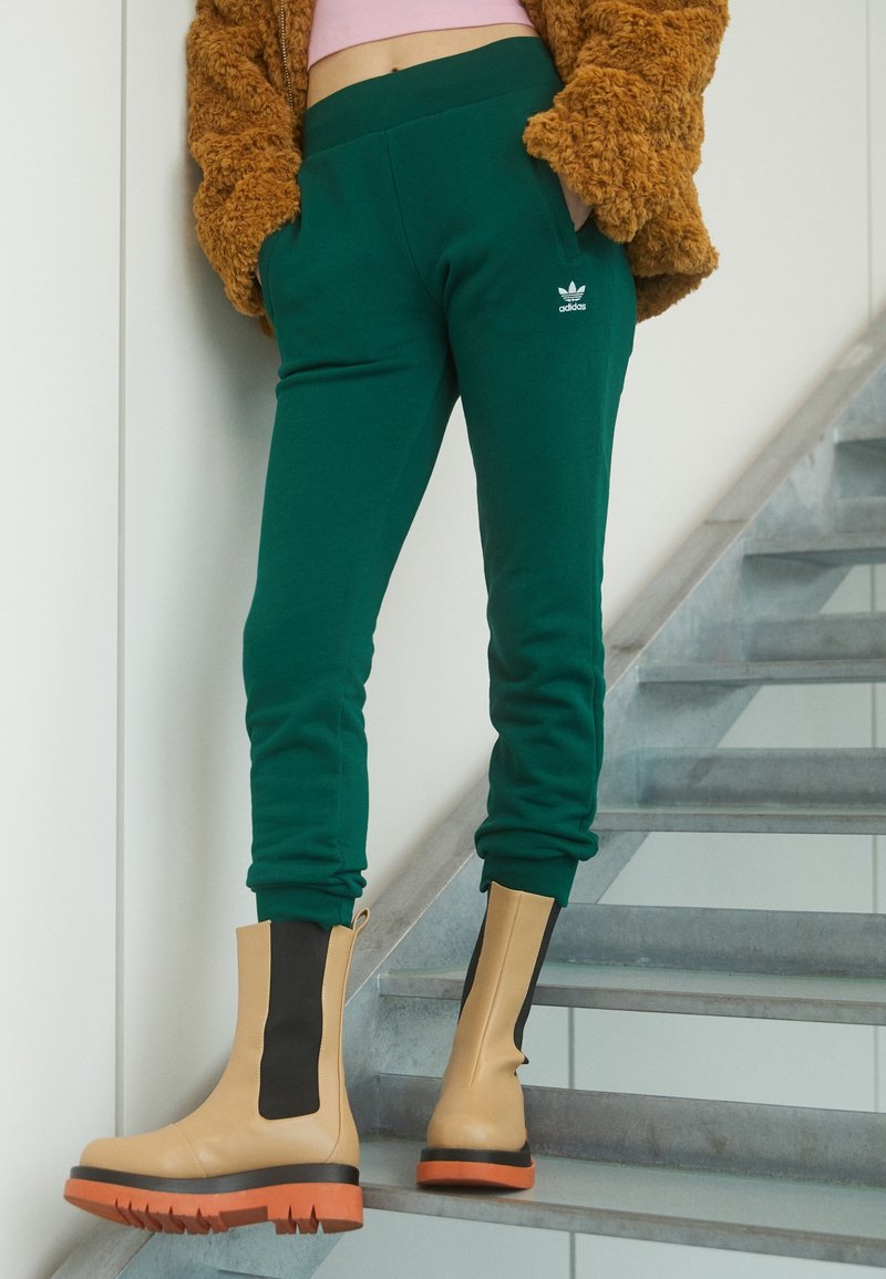 Zelené tepláky s bočními kapsami, s logem Adidas. Spárované s béžovými kotníkovými botami s černými akcenty a oranžovými podrážkami na schodech.