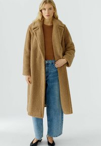 Vrouw in een lange bruine teddyjas over een bruine trui, met blauwe wijde jeans en zwarte schoenen, staand tegen een eenvoudige achtergrond.