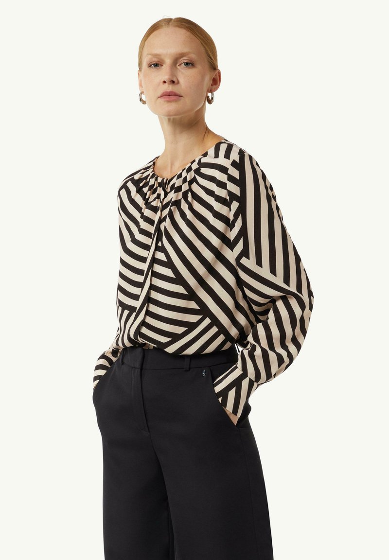 Schwarz-beige gestreifte Bluse mit langen Ärmeln und gerafftem Ausschnitt. Kombiniert mit hochgeschnittenen schwarzen Hosen, die eine lockere Silhouette erzeugen.