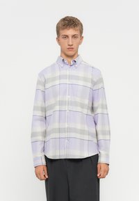 Joven vistiendo una camisa de cuadros en tonos púrpura pastel y gris, con pantalones negros, de pie contra un fondo blanco liso.