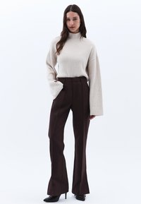 Beige geribbelde trui met hoge hals en brede mouwen, gecombineerd met bruin flair-pantalon. Zwarte hakken maken de look af.