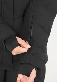 Der schwarze Puffer-Jacke Ärmel verfügt über eine Reißverschlusstasche, elastische Bündchen und eine strukturierte Oberfläche für Wärme und Komfort.