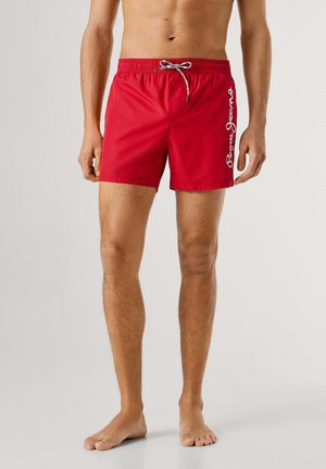 Modello maschile che indossa pantaloncini da bagno rossi con coulisse regolabile bianca e nera e logo bianco "Pepe Jeans" sulla coscia sinistra.
