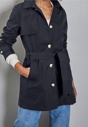 Person iført en sort trenchcoat med bælte og hvide knapper, hvid langærmet sweater under og lyseblå jeans, hånd i lommen.
