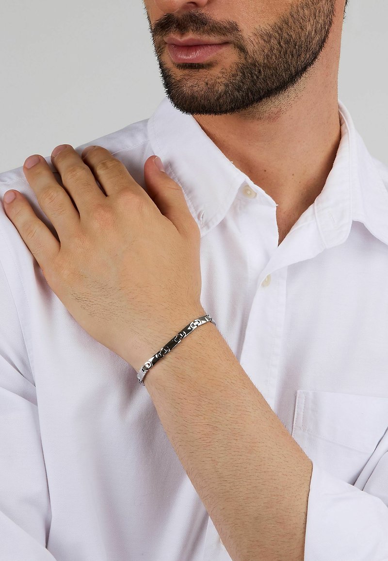 Bracciale in metallo con una finitura argento lucido, dettagli neri e elementi di design intricati, indossato su un polso marrone chiaro.