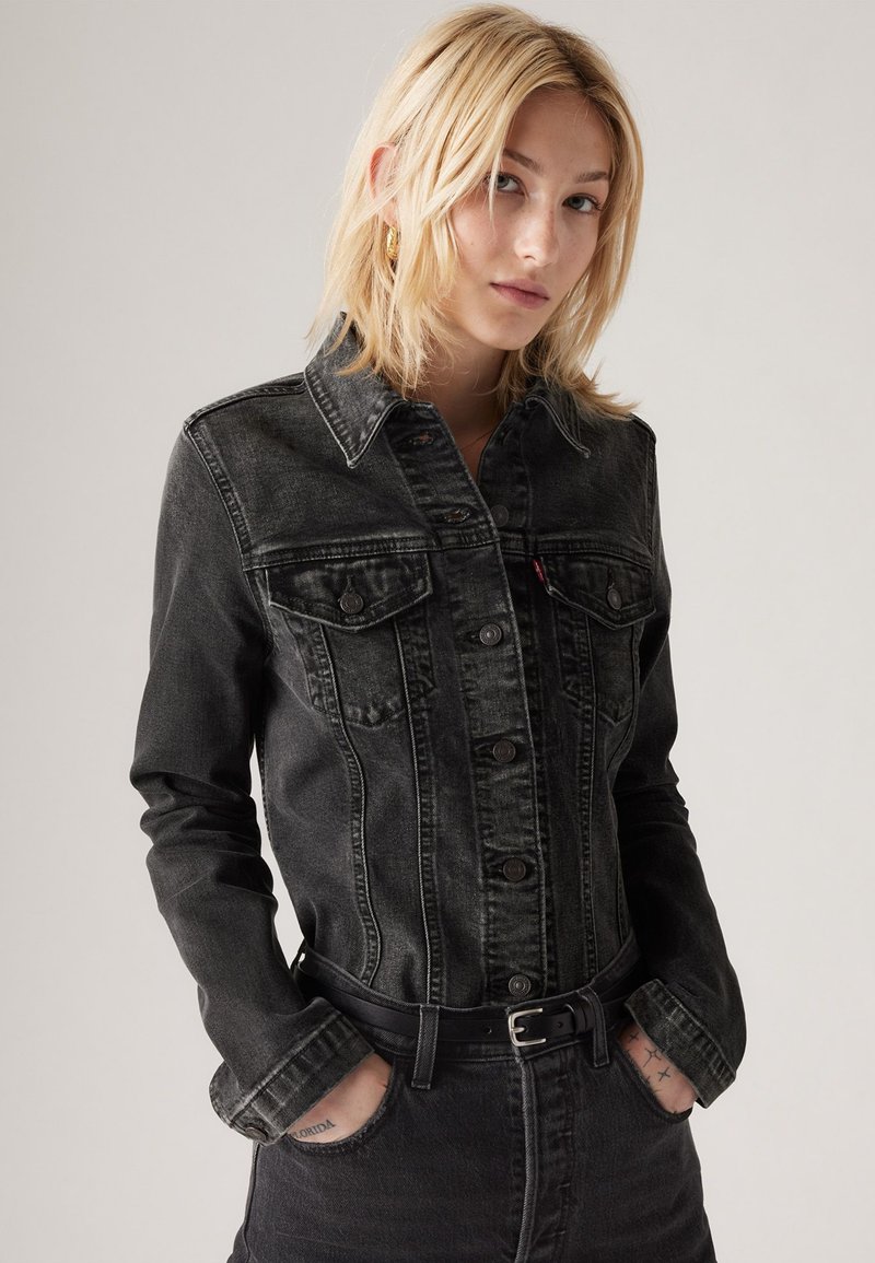Junge Frau mit blonden Haaren, die eine schwarze Denimjacke und passende schwarze Jeans trägt, mit Händen in den Taschen vor einem neutralen Hintergrund.