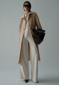 Trench coat bej cu accente de perle, cămașă albă cu guler, pantaloni albi cu croială largă și o geantă geometrică neagră. Accesoriile includ ochelari de soare închiși la culoare.
