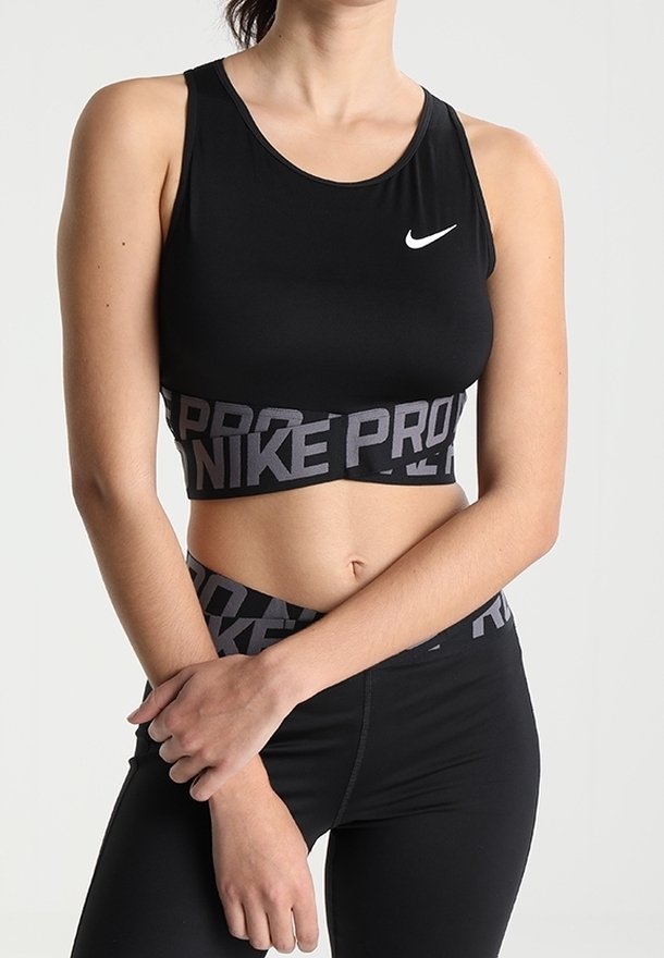 Žena v černém sportovním podprsence Nike Pro a legínách, stojící s rukama zkříženýma před jednoduchým světlým pozadím.