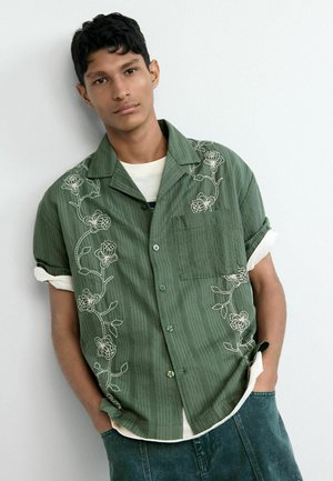 Jonge man met kort krullend haar, gekleed in een groen overhemd met korte mouwen en witte bloemenborduursels, staand met handen in zijn zakken.