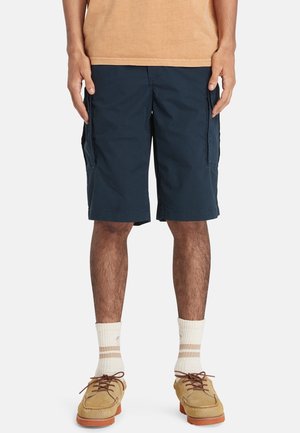 Marineblaue Cargo-Shorts mit mehreren Seitentaschen, aus leichtem Stoff gefertigt, mit entspannter Passform. Kombiniert mit beiger Fußbekleidung und Socken.