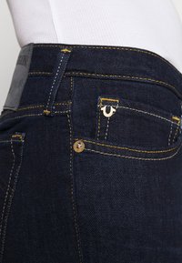 Los jeans de mezclilla azul oscuro presentan costuras amarillas, un pequeño detalle decorativo de logo y un parche de cuero genuino en la cinturilla.