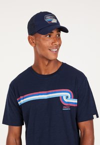 Camiseta azul marino con un diseño gráfico que presenta tres franjas horizontales en rojo, azul y azul claro, combinada con una gorra azul marina.