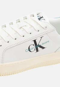 Witte sneaker met een gladde synthetische bovenkant, voorzien van een zwart "CK"-logo en turquoise "Calvin Klein Jeans"-tekst, en een gestructureerde crèmekleurige rubberen zool.