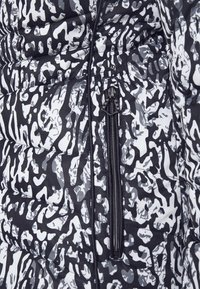 Doudoune avec un motif abstrait noir et blanc, texture matelassée, poche zippée et tissu lisse. Détail d'attache de fermeture éclair visible.