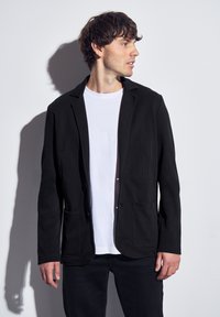 Svart kavaj i texturerat tyg, med slagkrage, två fickor fram och knappstängning framtill, stylad över en vit t-shirt.