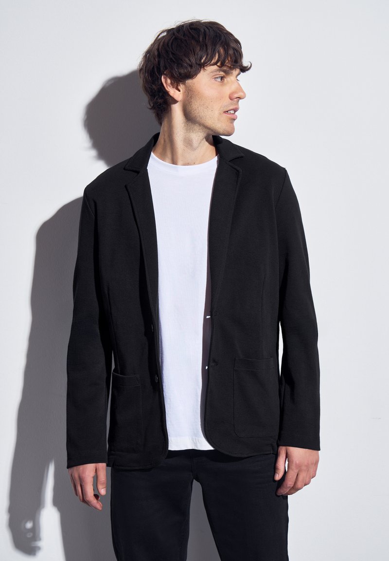 Svart kavaj i texturerat tyg, med slagkrage, två fickor fram och knappstängning framtill, stylad över en vit t-shirt.