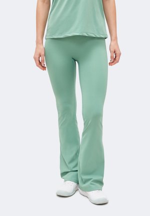 Άτομο που φορά μέντα πράσινο καμπάνα παντελόνι, ταιριαστό τοπ και λευκά αθλητικά παπούτσια, στέκεται μπροστά σε απλό ανοιχτό φόντο.