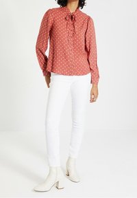 Rostfärgad blus med prickar, puffärmar, knytkrage och knappstängning, i kombination med vita skinny jeans och vita klackade ankelstövlar.
