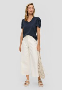 Marineblauw V-hals T-shirt met geplooide korte mouwen, gecombineerd met witte wijde broek en beige platform sandalen. Houdt een gevlochten tas vast.