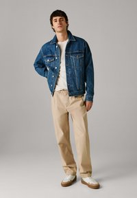 Ung man stående klädd i blå jeansjacka, vit t-shirt, beige byxor och vita sneakers mot en enkel grå bakgrund.
