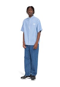 Una camicia a maniche corte con bottoni presenta righe verticali blu e bianche, abbinata a jeans in denim blu morbidi e sneakers nere.
