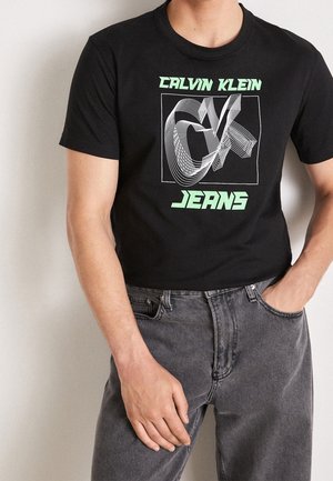 Czarna bawełniana koszulka z grafiką i białymi oraz neonowo-zielonymi napisami. W zestawie z szarymi dżinsami. Krótkie rękawy i okrągły dekolt.