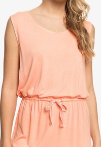 Robe sans manches de couleur pêche avec un décolleté rond, taille cintrée et détails à cordon. Fabriquée dans un tissu doux et léger avec une texture lisse.