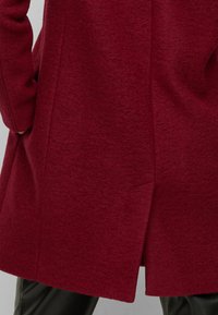 Rode wollen jas met een gestructureerde afwerking, zijzakken en een rugsplit voor bewegingsgemak. Het ontwerp is getailleerd en heeft lange mouwen.