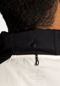 Col de la veste en noir avec une fermeture à bouton, présentant un panneau en tissu blanc en dessous. L'étiquette de la veste est visible sur le dessous.