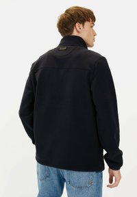 Marineblaues Fleece-Jacket mit hohem Kragen, glatter Textur, subtilen Nähdetails und einem Patch-Label auf der Rückseite, kombiniert mit hellen Jeans.