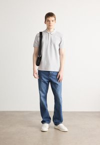 Polo gris avec un col, associé à un jean en denim bleu. Jeune modèle masculin tient un sac à épaule noir, portant des baskets blanches. Fond simple.