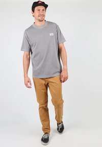 T-shirt gris à manches courtes avec un logo en patch, associé à un chino beige et des baskets noires. L'ensemble présente une coupe décontractée et des lignes épurées.