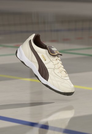 Λευκό και καφέ αθλητικό παπούτσι Puma με ορατά κορδόνια και λογότυπο, αιωρούμενο πάνω από το δάπεδο ενός γυμναστηρίου με πολύχρωμες γραμμές γηπέδου.