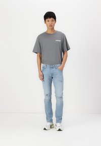Grå t-skjorte med "Wrangler"-logo, kombinert med lyseblå ripped jeans og hvite joggesko. Modellen står foran en klar hvit bakgrunn.