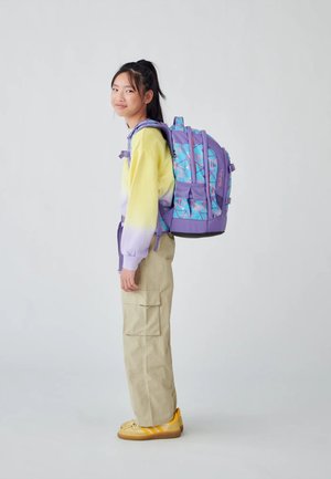 Backpack in purple with blue geometric patterns; features adjustable straps, multiple pockets, and a sturdy base. Worn by a person standing.