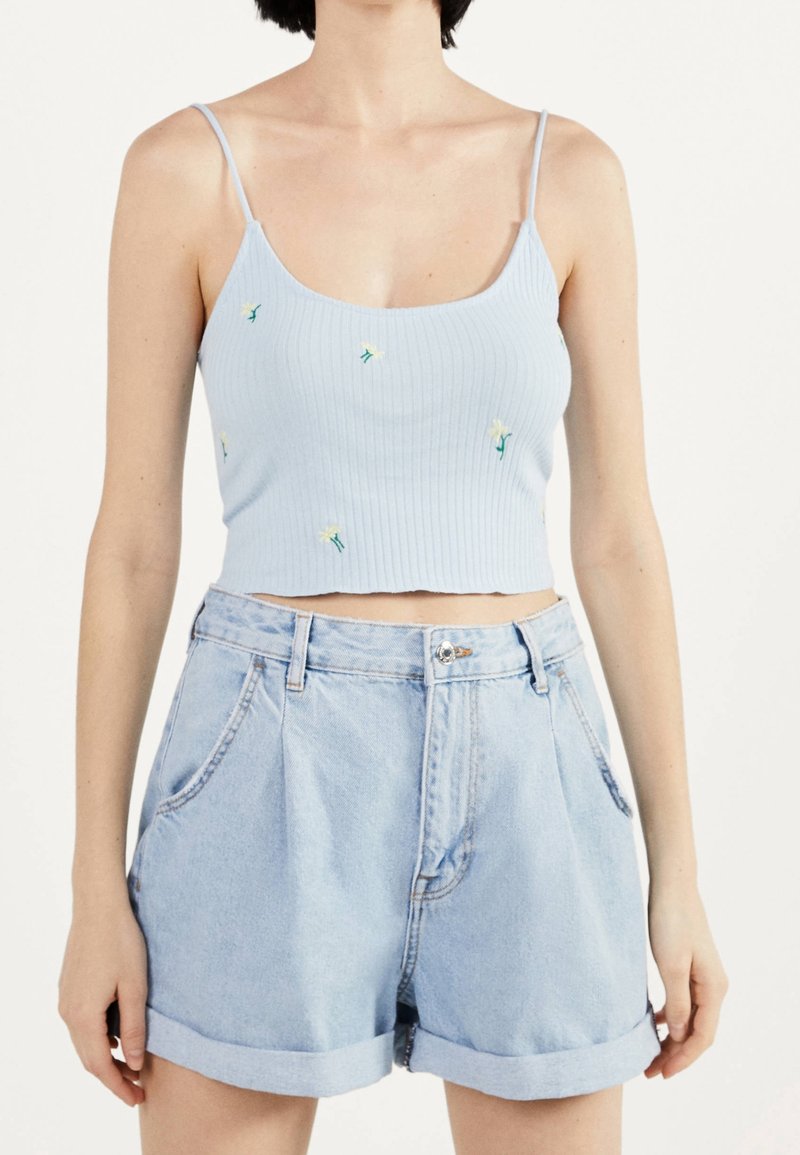 Kvinna som bär en ljusblå ribbad crop top med små vita blommor och högmidjade denimshorts med uppvikta fållar, stående mot en enfärgad bakgrund.
