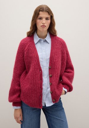 Vrouw in een rode chunky gebreide cardigan boven een blauw-wit gestreepte button-up shirt en blauwe spijkerbroek, staand met één hand in de zak.