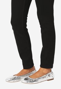 Stopy w srebrnych balerinach z motywem skóry węża w poł ączeniu z czarnymi dopasowanymi spodniami na białym tle.