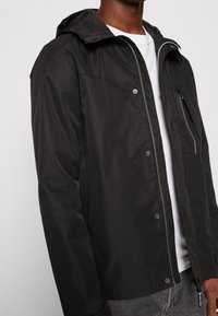 Zwarte waterdichte jas met een capuchon, ritssluiting aan de voorkant en drukknopen, twee zijzakken en een borstzak met een verticale ritssluiting.