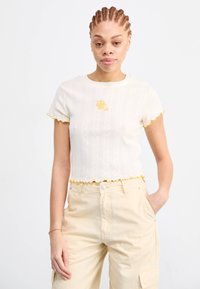 Crème gekort t-shirt met gele gegolfde randen en bloem borduursel, met een verticale ribbelpatroon en korte mouwen.