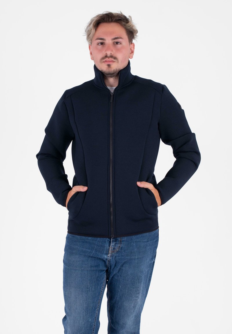 Giacca zip-up blu navy con colletto alto, realizzata in tessuto testurizzato. Presenta due tasche laterali e un design aderente. Indossata con jeans blu.