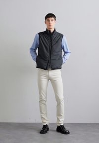 Black padded vest over a light blue shirt, cream trousers, and black shoes. Simple design, front zipper, and pockets.
