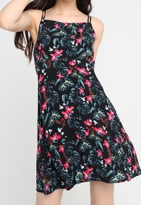 Kobieta w bezrękawnej czarnej sukience z różowym, niebieskim i zielonym tropikalnym wzorem kwiatowym, stojąca na tle jednolitego jasnego tła.