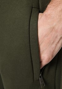 Tkanina joggerów w oliwkowym zielonym kolorze, z zbliżeniem na boczną kieszeń i widocznym detalem zamka. Gładka faktura, dopasowany krój, minimalistyczny design.