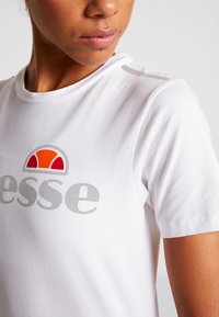 Hvid kortærmet t-shirt lavet af bomuld. Har et stort gråt logo med røde og orange accenter. Glat tekstur og rund halsudskæring.